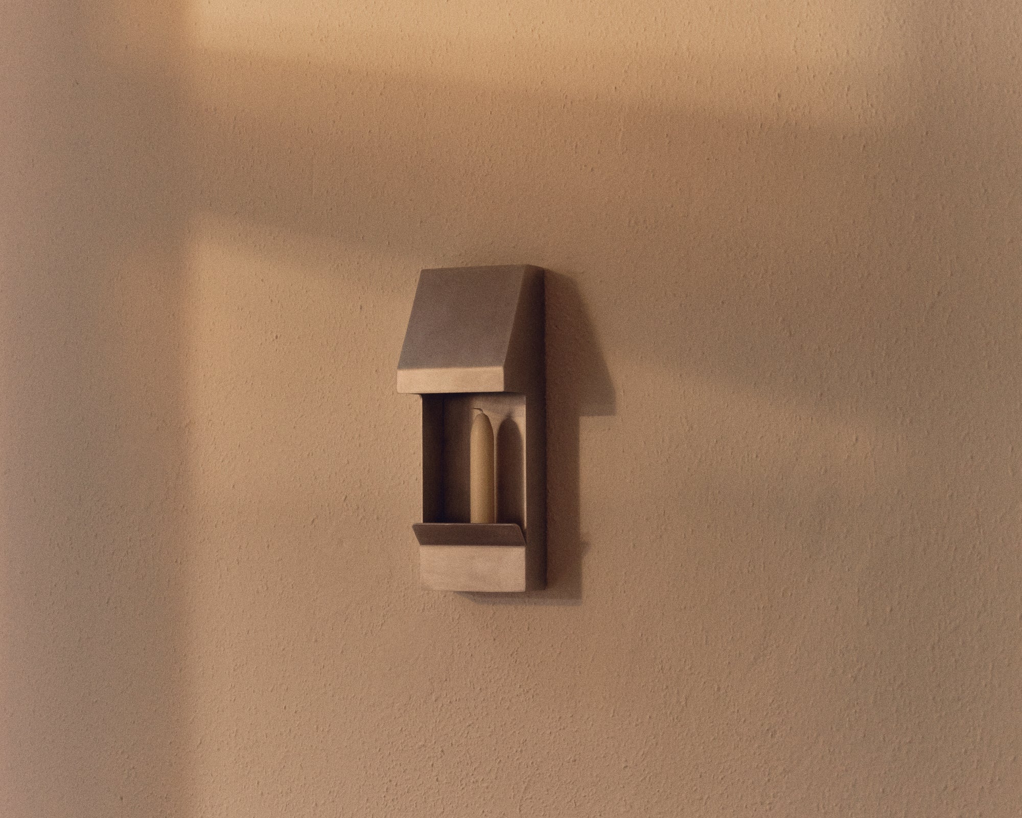 this aluminium tapered candle sconce is affixed to an ivory wall, which is bathed in afternoon sunlight. The aluminium sconce houses an ivory tapered candle, which is not illuminated but creates light and shadow on the wall. The sconce is a perfect addition for interior stylists, decorators, those seeking to elevate their interior and add warmth and natural illumination to their space.